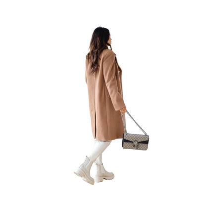 Woman in a beige coat holding a handbag in a minimalistic room.
