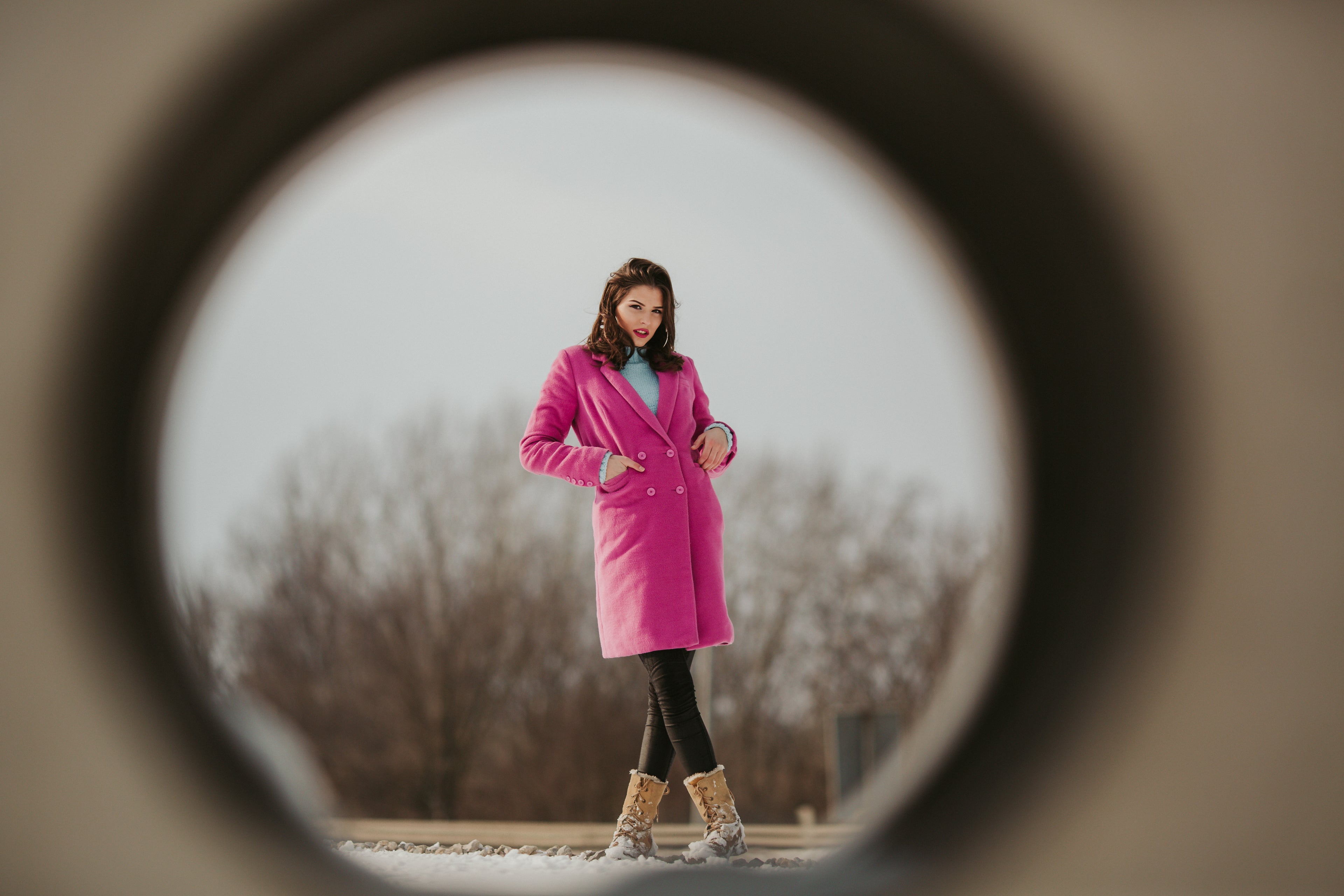Woman in a pink coat standing outdoors, framed by a circular lens view.